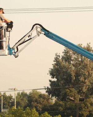 Man in a cherry picker operating machinery outdoors, with trees and equipment.